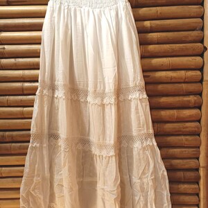 May include: A white, tiered skirt with a smocked waistband and lace trim. The skirt is made of a lightweight fabric and hangs from a black hanger. The skirt is displayed against a bamboo background.