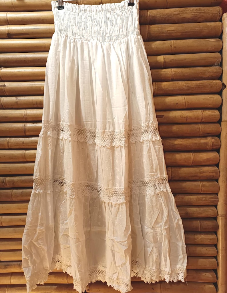 May include: A white, tiered skirt with a smocked waistband and lace trim. The skirt is made of a lightweight fabric and hangs from a black hanger. The skirt is displayed against a bamboo background.