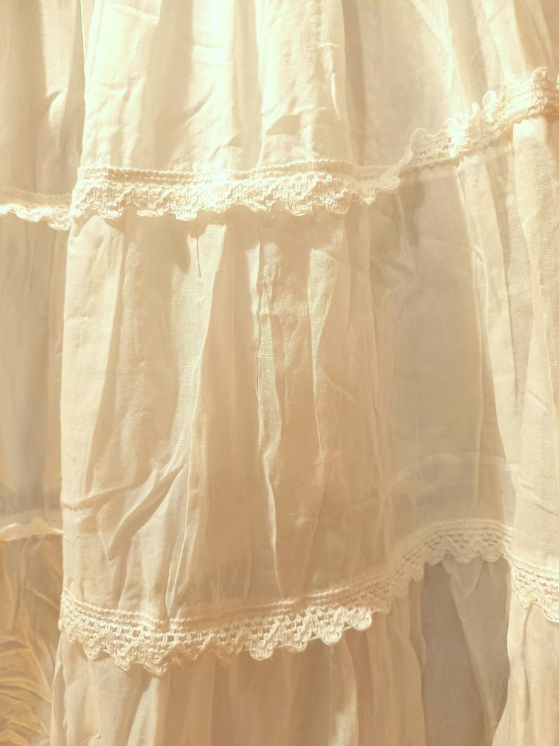 May include: Close-up of a white, layered fabric with lace detailing. The fabric is light and flowing, with visible creases and shadows. The lace trim adds a delicate touch, creating a soft, romantic aesthetic. The image suggests a garment or textile.