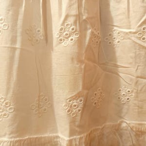 May include: Close-up of off-white fabric with embroidered floral eyelet patterns. The fabric has a ruffled hem and is slightly creased, suggesting it is part of a garment or home decor item.