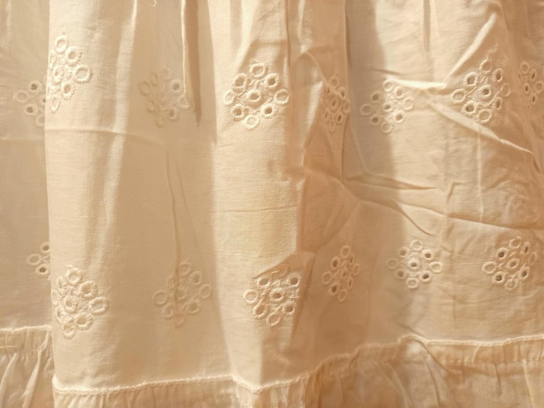 May include: Close-up of off-white fabric with embroidered floral eyelet patterns. The fabric has a ruffled hem and is slightly creased, suggesting it is part of a garment or home decor item.