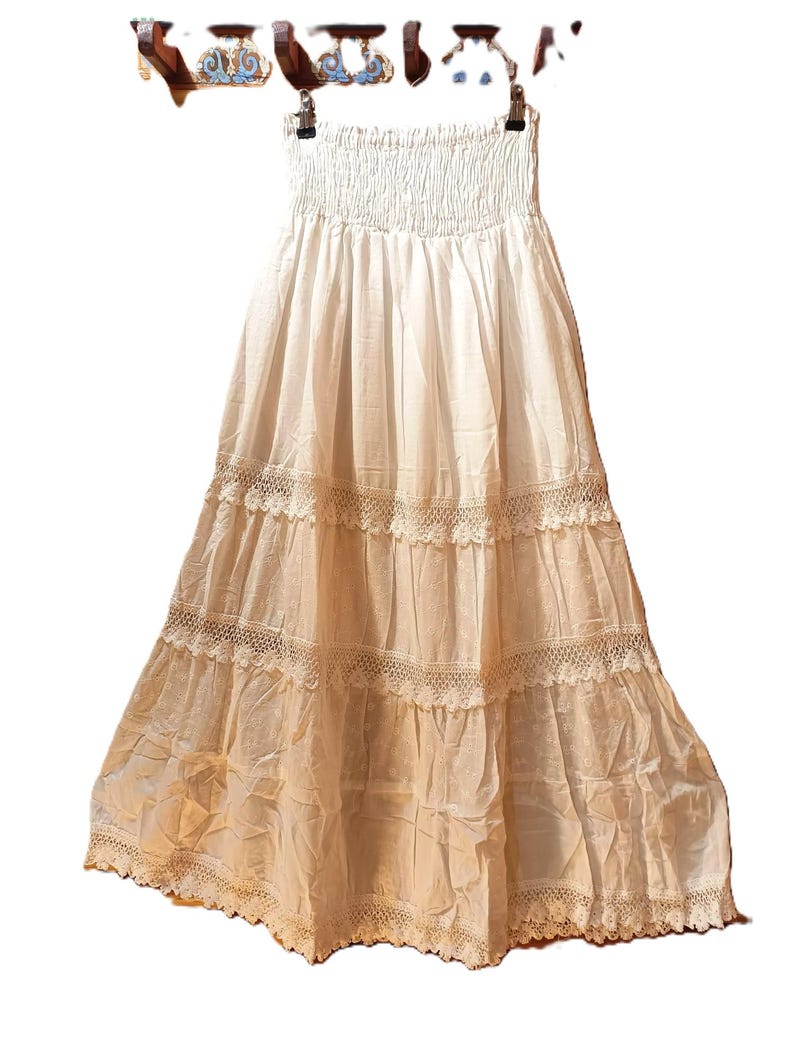 May include: A white, strapless maxi skirt with a smocked bodice and tiered, lace-trimmed layers. The skirt is hanging on a hanger. The fabric appears to be lightweight and flowing, suitable for warm weather. The skirt has a bohemian style.
