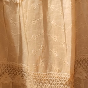 May include: Close-up of a cream-coloured fabric with intricate embroidery and lace detailing. The fabric features delicate floral patterns and openwork designs, creating a textured and layered appearance. The overall aesthetic suggests a vintage or handcrafted textile.