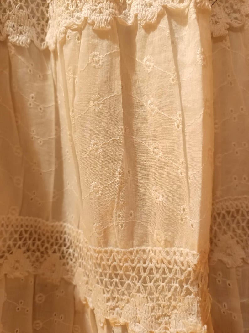 May include: Close-up of a cream-coloured fabric with intricate embroidery and lace detailing. The fabric features delicate floral patterns and openwork designs, creating a textured and layered appearance. The overall aesthetic suggests a vintage or handcrafted textile.