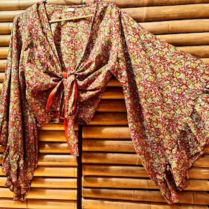 May include: A red, yellow, and green patterned top with long, flared sleeves. The top has a V-neck and a tie closure at the waist. The fabric has a floral design. The top is hanging on a wooden hanger.