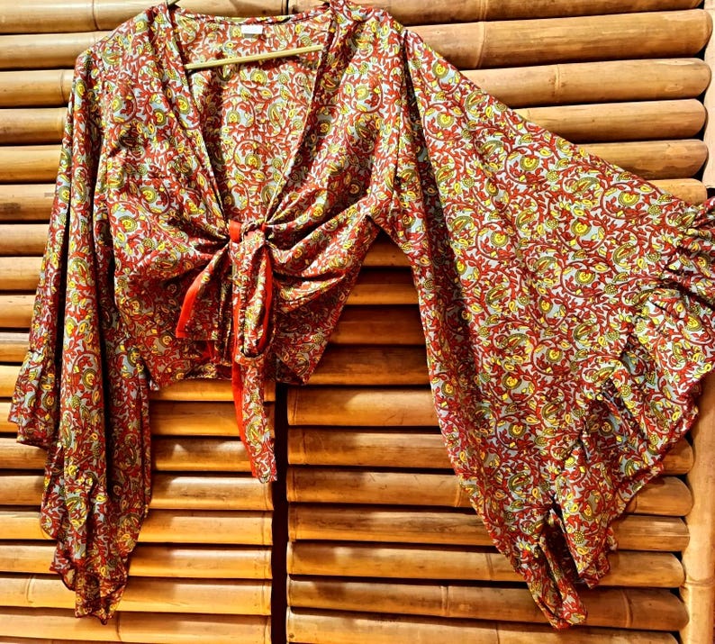 May include: A red, yellow, and green patterned top with long, flared sleeves. The top has a V-neck and a tie closure at the waist. The fabric has a floral design. The top is hanging on a wooden hanger.