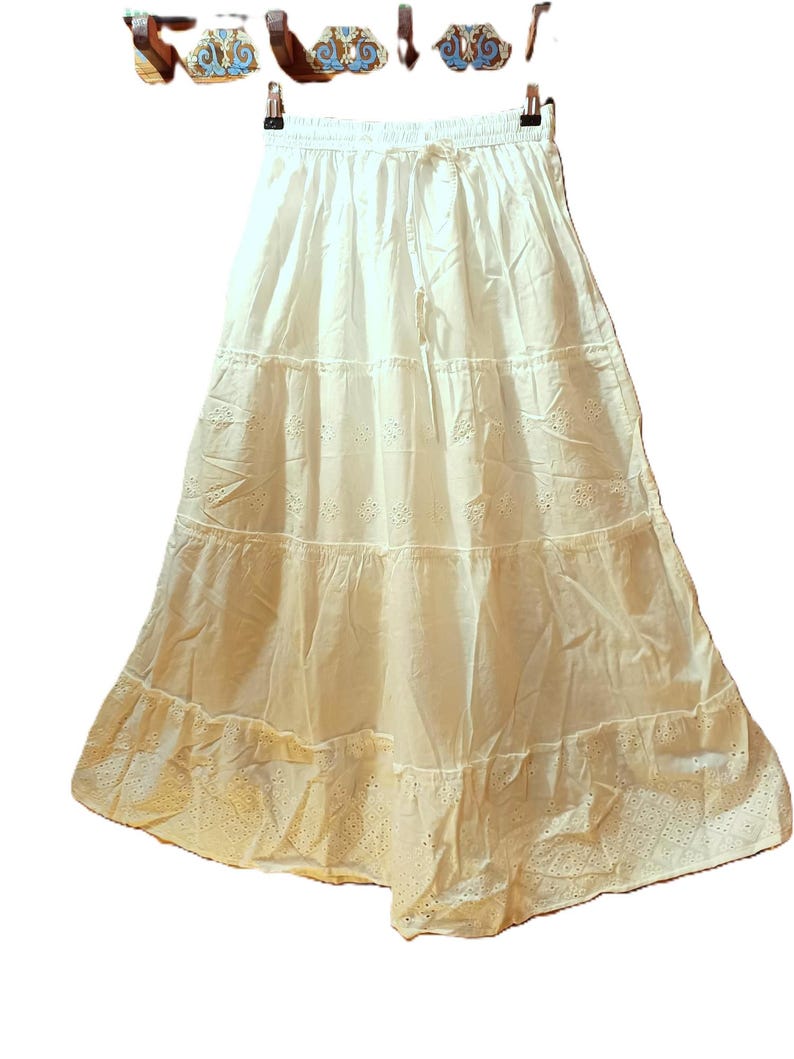 May include: A white tiered skirt with a drawstring waist. The skirt has eyelet detailing on the bottom tier and a flowing, bohemian style. The skirt is hanging on a hanger against a white background.