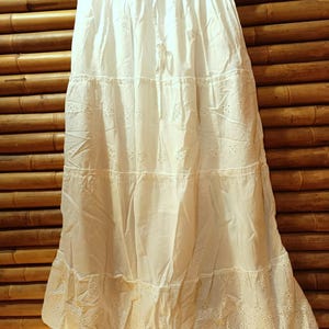 May include: A white tiered skirt with eyelet detailing and a drawstring waist. The skirt is made of a lightweight fabric and hangs from a black hanger against a bamboo background. The skirt is approximately 36 inches long.