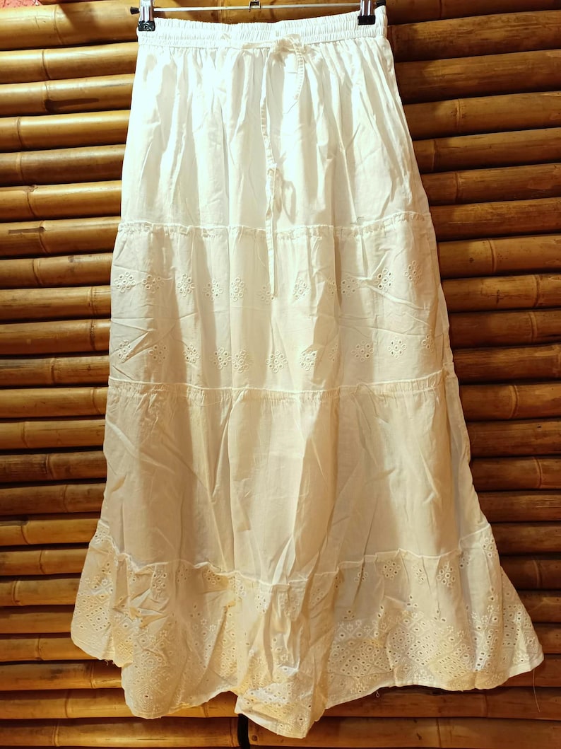 May include: A white tiered skirt with eyelet detailing and a drawstring waist. The skirt is made of a lightweight fabric and hangs from a black hanger against a bamboo background. The skirt is approximately 36 inches long.