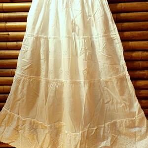 May include: A white tiered skirt with a drawstring waist hangs from a black hanger. The skirt features eyelet embroidery and a ruffled hem. The fabric appears to be lightweight cotton. The skirt is approximately 30 inches long.