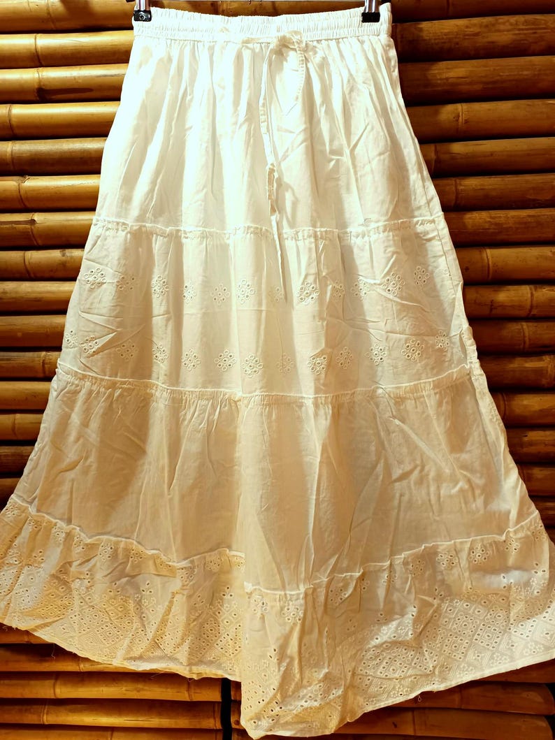 May include: A white tiered skirt with a drawstring waist hangs from a black hanger. The skirt features eyelet embroidery and a ruffled hem. The fabric appears to be lightweight cotton. The skirt is approximately 30 inches long.