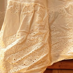 May include: Close-up of off-white fabric with intricate eyelet embroidery. The fabric features detailed patterns of circles, diamonds, and floral designs, creating a textured and delicate appearance. The light color suggests a summer garment.