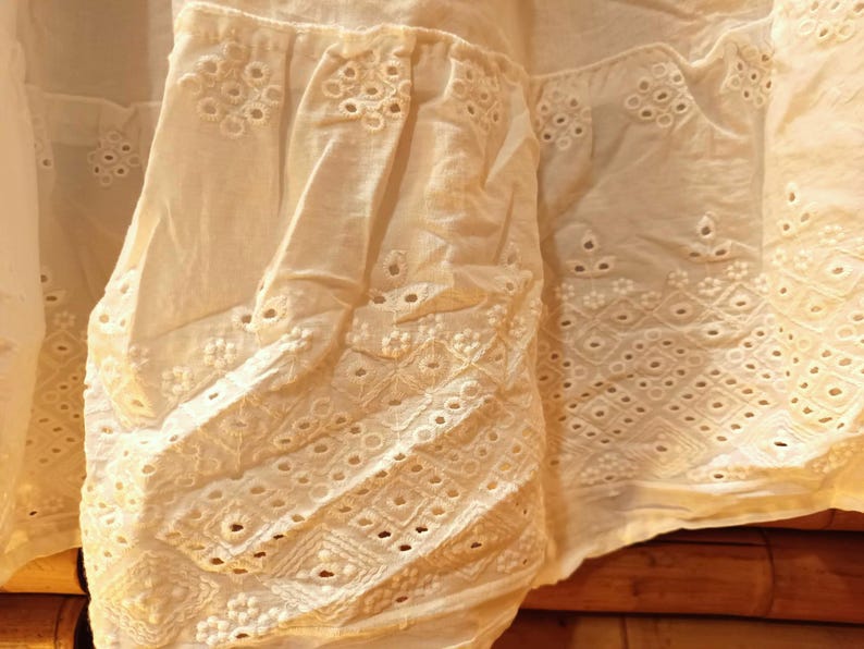May include: Close-up of off-white fabric with intricate eyelet embroidery. The fabric features detailed patterns of circles, diamonds, and floral designs, creating a textured and delicate appearance. The light color suggests a summer garment.