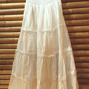 May include: A white, tiered maxi skirt with a smocked waistband and lace trim. The skirt is made of a lightweight fabric and hangs from a metal hanger against a bamboo background. The skirt is a neutral color.