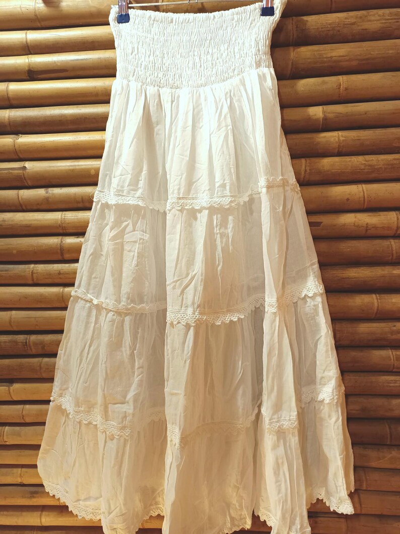 May include: A white, tiered maxi skirt with a smocked waistband and lace trim. The skirt is made of a lightweight fabric and hangs from a metal hanger against a bamboo background. The skirt is a neutral color.