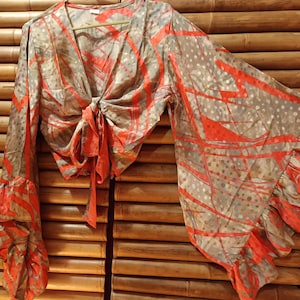 May include: A tie-front blouse with long, flared sleeves. The blouse features a V-neck and a tie closure at the waist. The fabric has a gray background with a red abstract pattern and polka dots. The sleeves have a ruffled hem.