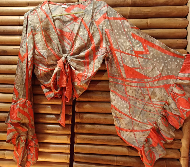 May include: A tie-front blouse with long, flared sleeves. The blouse features a V-neck and a tie closure at the waist. The fabric has a gray background with a red abstract pattern and polka dots. The sleeves have a ruffled hem.