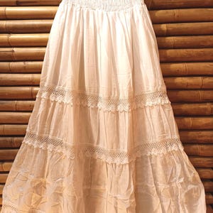 May include: A white, tiered maxi skirt with a smocked waistband and lace trim. The skirt is made of a lightweight fabric and hangs from a black hanger. The skirt is approximately 91 cm long.