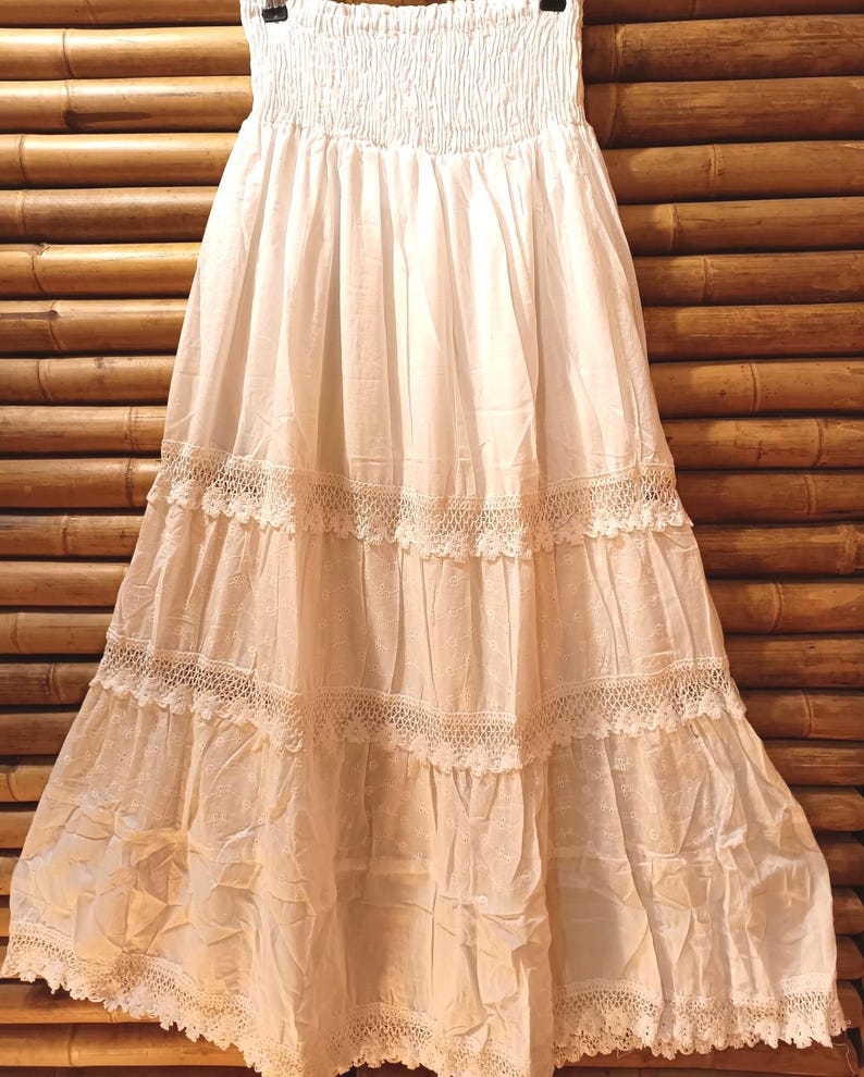 May include: A white, tiered maxi skirt with a smocked waistband and lace trim. The skirt is made of a lightweight fabric and hangs from a black hanger. The skirt is approximately 91 cm long.