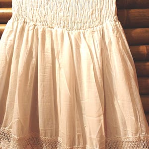 May include: Off-white skirt with a smocked bodice and a flowing, tiered design. The skirt features a delicate, crocheted lace trim along the hemline. The fabric appears lightweight and airy, suitable for warm weather.