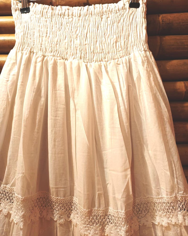 May include: Off-white skirt with a smocked bodice and a flowing, tiered design. The skirt features a delicate, crocheted lace trim along the hemline. The fabric appears lightweight and airy, suitable for warm weather.