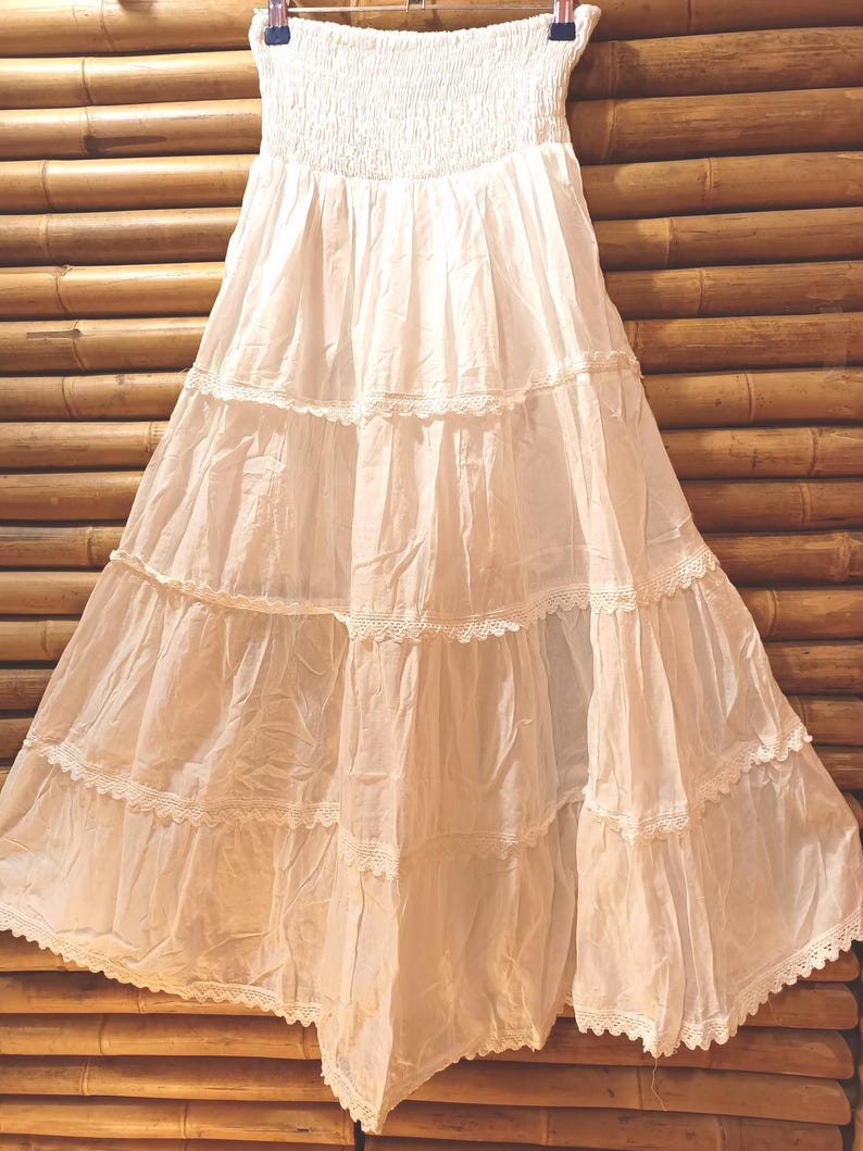 May include: A white, tiered maxi skirt with a smocked bodice and lace trim. The skirt is made of a lightweight fabric and hangs from a hanger against a bamboo background. The skirt has a bohemian style.
