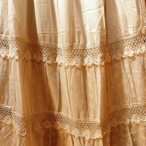 May include: Close-up of a cream-coloured skirt with multiple tiers. Each tier is adorned with intricate lace trim. The fabric has a delicate, slightly sheer quality, with subtle embroidered details adding texture and visual interest.