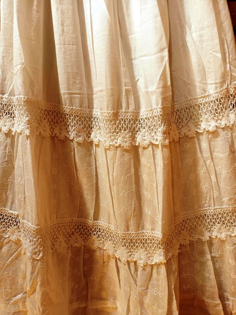 May include: Close-up of a cream-coloured skirt with multiple tiers. Each tier is adorned with intricate lace trim. The fabric has a delicate, slightly sheer quality, with subtle embroidered details adding texture and visual interest.