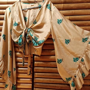 May include: A beige tie-front blouse with long, flared sleeves and a ruffled hem. The blouse is adorned with a teal floral pattern. The top is hanging on a hanger.