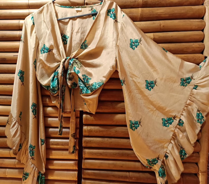 May include: A beige tie-front blouse with long, flared sleeves and a ruffled hem. The blouse is adorned with a teal floral pattern. The top is hanging on a hanger.