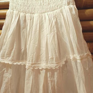 May include: A white tiered skirt with a smocked bodice. The skirt has delicate lace trim along the tiers, creating a flowing silhouette. The fabric appears lightweight and airy, suitable for warmer weather.