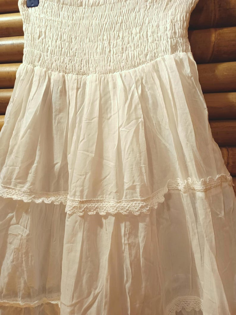 May include: A white tiered skirt with a smocked bodice. The skirt has delicate lace trim along the tiers, creating a flowing silhouette. The fabric appears lightweight and airy, suitable for warmer weather.