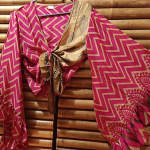 May include: A pink and gold wrap top with a chevron pattern and long, flared sleeves. The top is tied at the waist and features a contrasting beige panel with a leaf design. The sleeves have a ruffled hem.