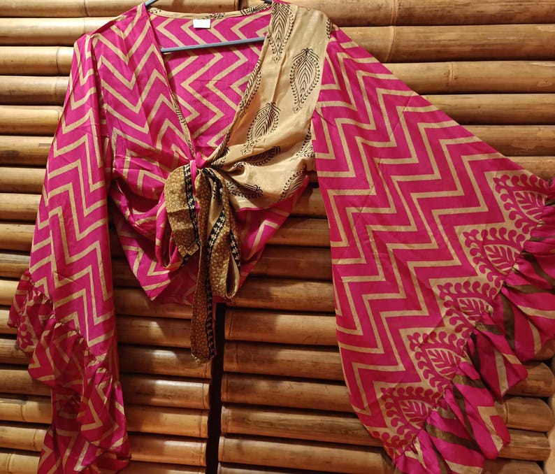 May include: A pink and gold wrap top with a chevron pattern and long, flared sleeves. The top is tied at the waist and features a contrasting beige panel with a leaf design. The sleeves have a ruffled hem.