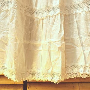 May include: Close-up of a white, tiered skirt with detailed eyelet and lace. The fabric is lightweight and slightly creased. The hemline features a delicate, scalloped lace trim, giving a vintage appearance. The skirt is made of a soft, breathable material.
