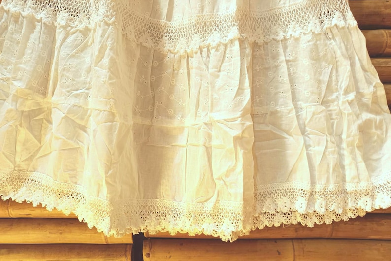 May include: Close-up of a white, tiered skirt with detailed eyelet and lace. The fabric is lightweight and slightly creased. The hemline features a delicate, scalloped lace trim, giving a vintage appearance. The skirt is made of a soft, breathable material.