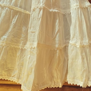 May include: A cream-coloured, tiered skirt with delicate lace trim. The skirt features multiple layers of fabric, creating a flowing and voluminous silhouette. The lace detailing adds a touch of elegance to the garment. The skirt is made of a lightweight material.