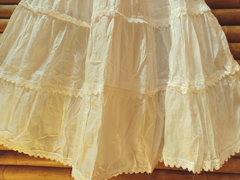 May include: A cream-coloured, tiered skirt with delicate lace trim. The skirt features multiple layers of fabric, creating a flowing and voluminous silhouette. The lace detailing adds a touch of elegance to the garment. The skirt is made of a lightweight material.