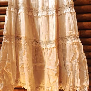 May include: An off-white tiered skirt featuring detailed lace and eyelet patterns. The skirt is composed of several layers, each embellished with delicate lace edging. The fabric seems light and flowing, possibly vintage or bohemian. The skirt is likely made of cotton or a similar natural fibre.