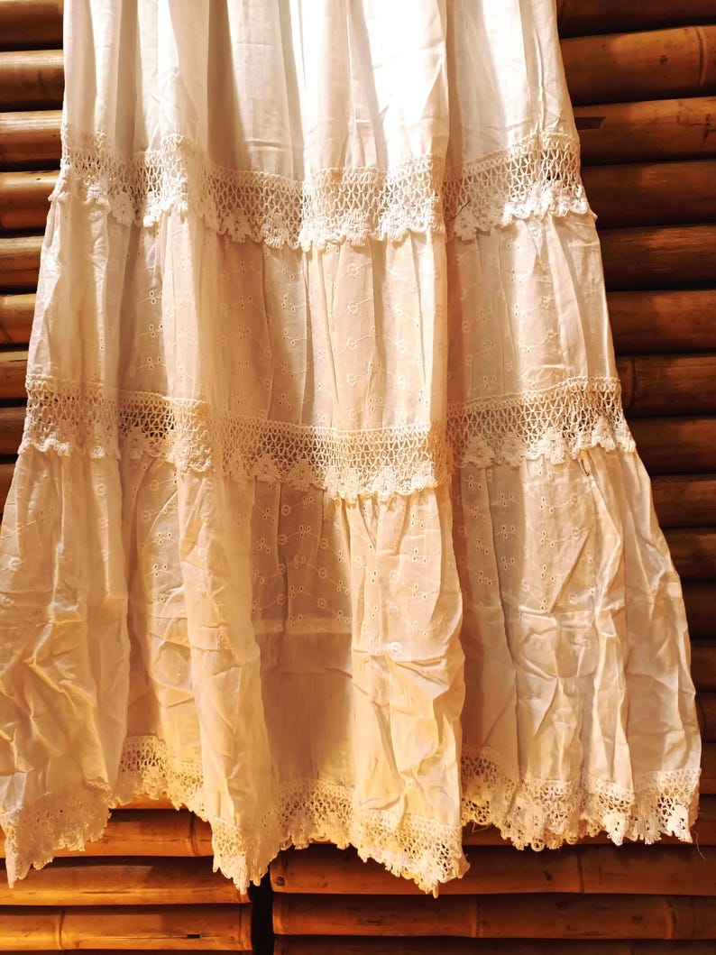 May include: An off-white tiered skirt featuring detailed lace and eyelet patterns. The skirt is composed of several layers, each embellished with delicate lace edging. The fabric seems light and flowing, possibly vintage or bohemian. The skirt is likely made of cotton or a similar natural fibre.