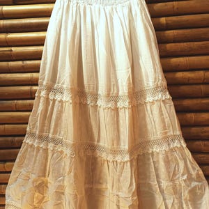 May include: A white, tiered skirt with a smocked waistband and lace trim. The skirt is made of a lightweight fabric and hangs from a black hanger against a bamboo background. The skirt has a bohemian style.