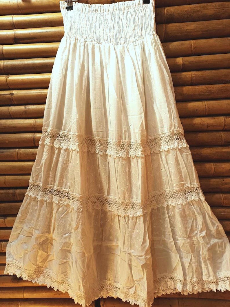 May include: A white, tiered skirt with a smocked waistband and lace trim. The skirt is made of a lightweight fabric and hangs from a black hanger against a bamboo background. The skirt has a bohemian style.