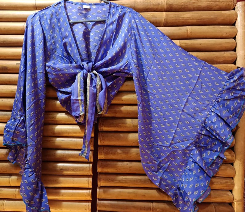 May include: A blue wrap top with a small, repeating pattern in yellow and white. The top has long, flared sleeves with ruffled cuffs and a tie closure at the waist. The fabric appears to be lightweight.