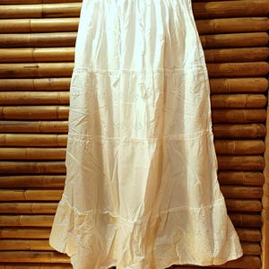 May include: A long, off-white tiered skirt with an elastic waistband. The skirt features eyelet detailing along the hemline and between the tiers. The skirt is hanging on a hanger against a bamboo background.