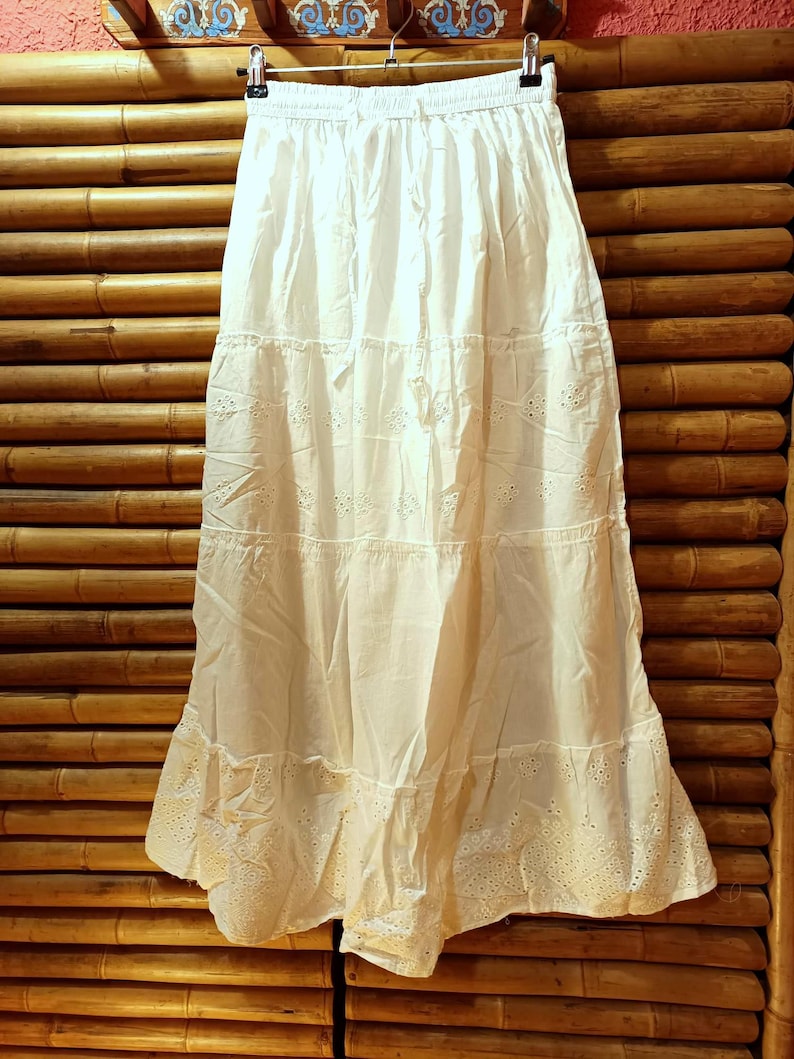 May include: A long, off-white tiered skirt with an elastic waistband. The skirt features eyelet detailing along the hemline and between the tiers. The skirt is hanging on a hanger against a bamboo background.