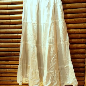 May include: A white tiered skirt with eyelet detailing, hanging on a metal hanger. The skirt has a drawstring waist and is made of a lightweight fabric. The skirt is displayed against a bamboo background.