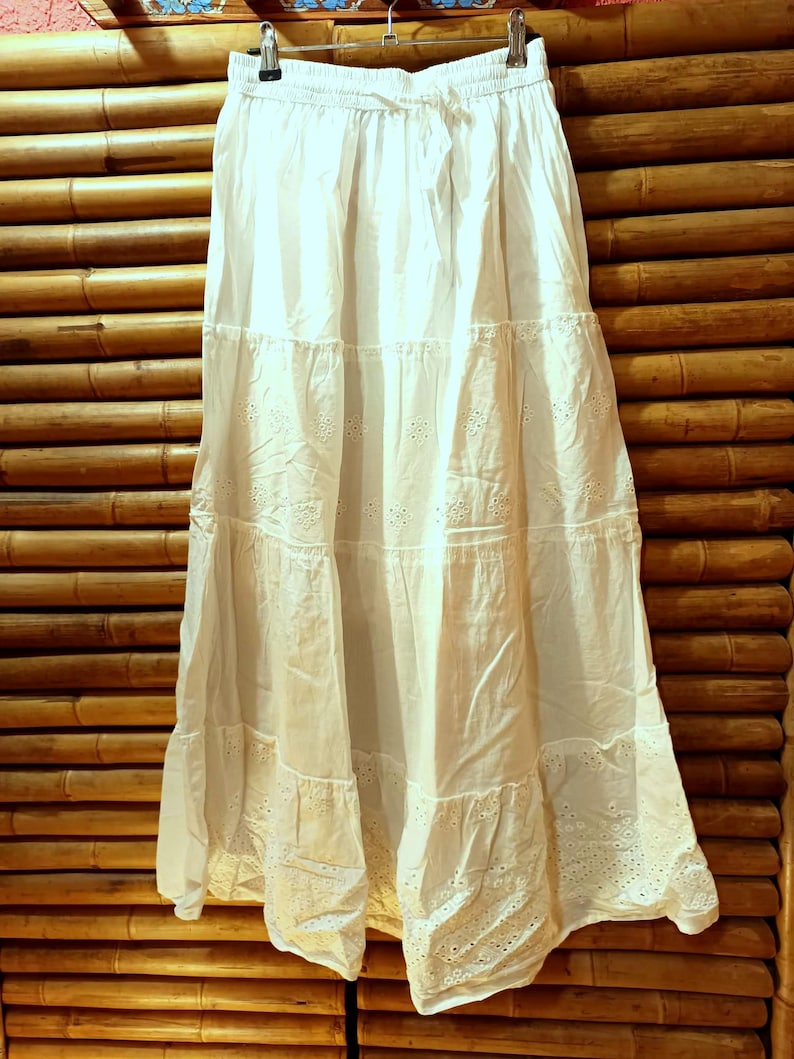 May include: A white tiered skirt with eyelet detailing, hanging on a metal hanger. The skirt has a drawstring waist and is made of a lightweight fabric. The skirt is displayed against a bamboo background.