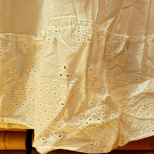 May include: Close-up of a cream-colored fabric with intricate eyelet embroidery. The fabric is layered, with a ruffled edge and detailed geometric and floral patterns. The texture is soft and delicate, suggesting a vintage or handmade item.