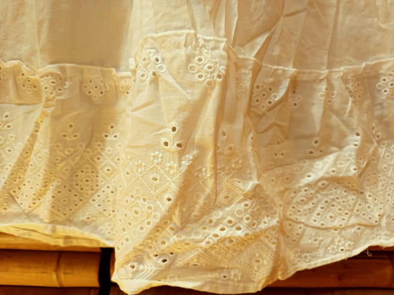 May include: Close-up of a cream-colored fabric with intricate eyelet embroidery. The fabric is layered, with a ruffled edge and detailed geometric and floral patterns. The texture is soft and delicate, suggesting a vintage or handmade item.
