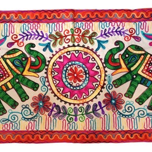 Può includere: Un arazzo rettangolare in tessuto con sfondo color crema e un vivace disegno ricamato. Il motivo include quattro elefanti in colori alternati viola e verde, un mandala circolare centrale e motivi floreali multicolori.